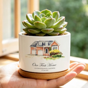 May include: A white ceramic planter with a wooden base containing a green succulent. The planter features a watercolor illustration of a house and the text "Our First Home", "4114 SEPULVEDA BLVD, CALIFORNIA", and "12th June 2026" printed on the front.