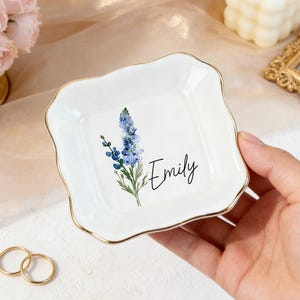 Könnte beinhalten: Ein weißes, quadratisches Schmuckschälchen mit goldenem Rand, mit einem blauen Blumendesign und dem Namen "Emily" in Schreibschrift. Zwei goldene Ringe sind im Vordergrund. Eine Kerze und ein Strauß rosa Rosen sind im Hintergrund.