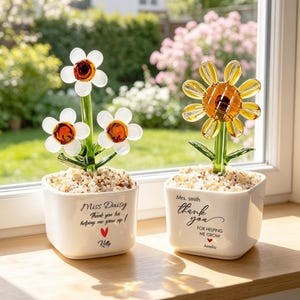 May include: Two white ceramic square planters filled with small stones, each containing a glass flower. The left planter says Miss Daisy, Thank you for helping me grow up, with a red heart and Kelly. The right says Mrs. Smith, thank you for helping me grow, with a red heart and Amelia.