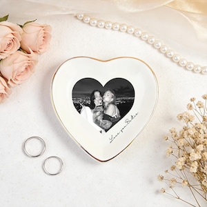 May include: A heart-shaped trinket dish with a gold rim, featuring a black and white photo of two people. The dish has the words "Love you Bestie" written on it. Two silver rings and flowers are also present.