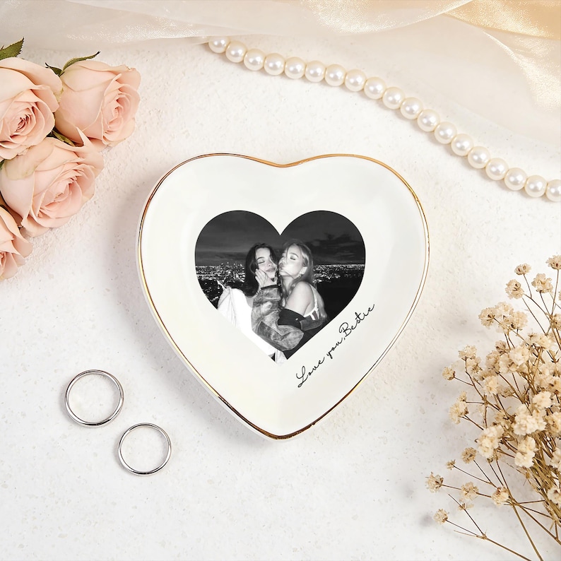 May include: A heart-shaped trinket dish with a gold rim, featuring a black and white photo of two people. The dish has the words "Love you Bestie" written on it. Two silver rings and flowers are also present.