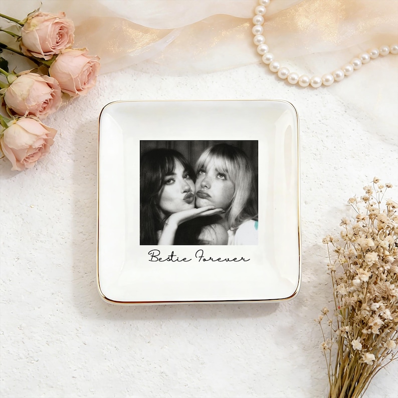 May include: Square white ceramic trinket dish with a gold rim. The center features a black and white photo of two women kissing, with the words "Bestie Forever" in script below. Decorative roses and pearls surround the dish.