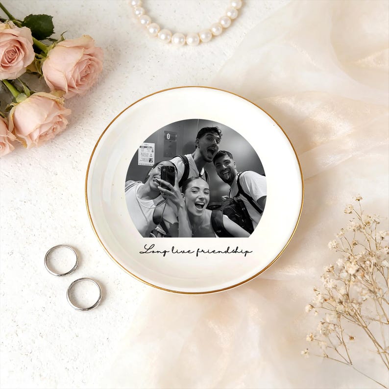 May include: A white ceramic trinket dish with a gold rim, featuring a black and white photo of four people. The dish has the phrase "Long live friendship" in script. Two silver rings and a pearl necklace are also visible.