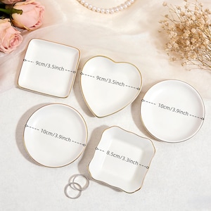 May include: Five white ceramic dishes with gold rims in various shapes: square (3.5 inches), heart (3.5 inches), round (3.9 inches), and a scalloped square (3.3 inches). Two silver rings are at the bottom. Decorative items.
