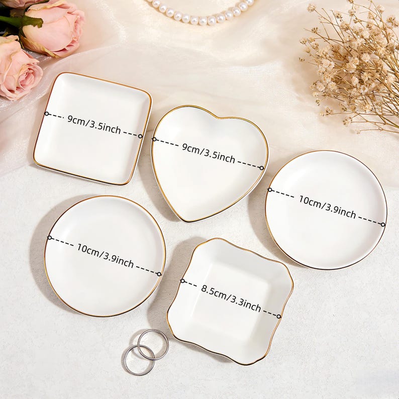 May include: Five white ceramic dishes with gold rims in various shapes: square (3.5 inches), heart (3.5 inches), round (3.9 inches), and a scalloped square (3.3 inches). Two silver rings are at the bottom. Decorative items.