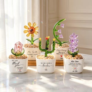 Può includere: Cinque statuette di piante in vetro in vasi di ceramica bianca riempiti di piccole pietre. Le piante includono una rosa rosa, un girasole giallo, un cactus verde, un mughetto viola e un grappolo di fiori viola, ognuno con testo inciso.