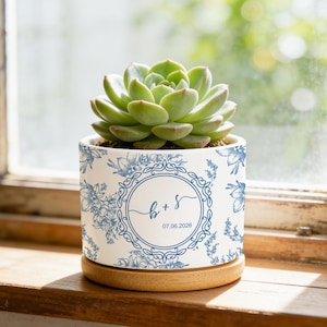 May include: A white ceramic cylindrical planter featuring a blue floral pattern and a central circular emblem with the letters b + s and the date 07.06.2026. The planter sits on a light-coloured wooden circular base and contains a green succulent.