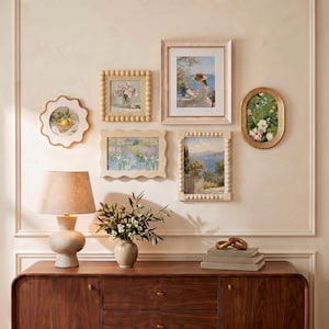 May include: A gallery wall arrangement featuring six framed art prints in various shapes, including scalloped, beaded, and oval frames. Below, a wooden sideboard holds a beige table lamp, a ceramic vase with greenery, and a stack of books.