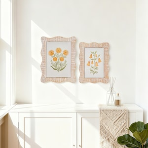 Könnte beinhalten: Zwei gerahmte Blumendrucke mit gewebten, wellenförmigen Rahmen. Ein Druck zeigt gelbe Blumen, der andere orangefarbene glockenförmige Blumen. Die Rahmen sind an einer weißen Wand über einem weißen Schrank angebracht. Ein Glasdiffusor und ein Makramee-Läufer sind ebenfalls sichtbar.