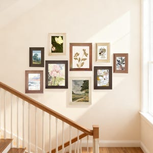 May include: A collection of framed art prints displayed on a wall. The prints feature various botanical and landscape scenes, including flowers, trees, and a butterfly. The frames are in various colors, including brown, gold, and white.
