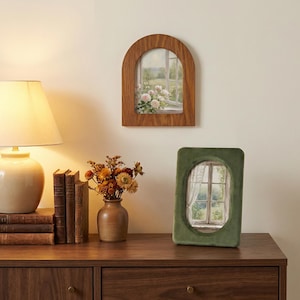May include: An arched wooden picture frame containing a print of a window with flowers, and an oval green velvet picture frame containing a print of a window with curtains, displayed alongside a ceramic vase of dried flowers and books.