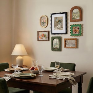 May include: A gallery wall arrangement featuring six framed artworks in various shapes and colors, including oval, rectangular, and arched frames. The wall art displays still life paintings of flowers, fruit, and window views, positioned above a wooden dining table.