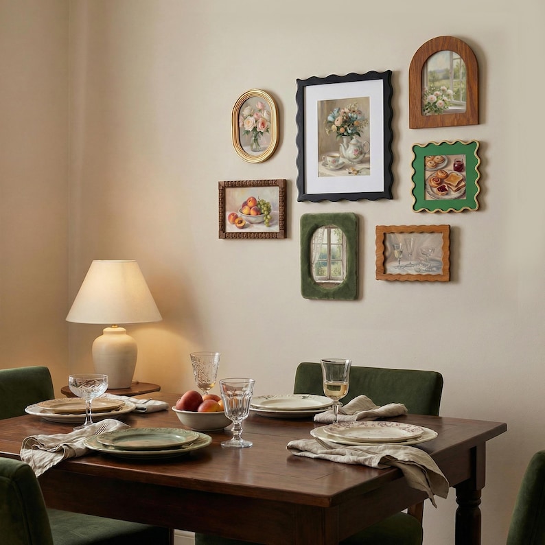 May include: A gallery wall arrangement featuring six framed artworks in various shapes and colors, including oval, rectangular, and arched frames. The wall art displays still life paintings of flowers, fruit, and window views, positioned above a wooden dining table.
