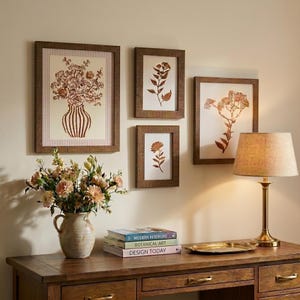 May include: Four framed botanical prints with brown wooden frames hang on a beige wall. The prints feature floral and plant illustrations in shades of brown and beige. A wooden desk holds a vase of flowers, books, and a lamp.