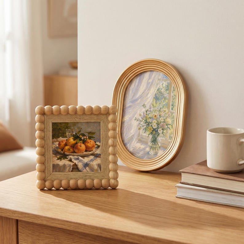 May include: A square wooden picture frame with a beaded border containing a still life print of peaches, next to an oval gold-toned ribbed picture frame displaying a print of a floral bouquet in a vase near a window with light blue curtains.