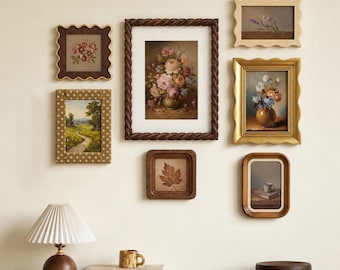 May include: A gallery wall arrangement featuring seven framed artworks, including floral paintings, a landscape, and a leaf study. Below, a pleated lamp, a textured mug, and two books labeled All Directions and Kerry Joyce sit on a surface.