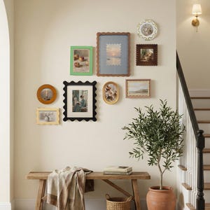 May include: A collection of framed artworks in various shapes and sizes, displayed on a wall. The frames are in diverse colours, including green, black, brown, and gold. The artworks depict landscapes, still lifes, and portraits. A wooden bench with a blanket and a potted plant are in the foreground.