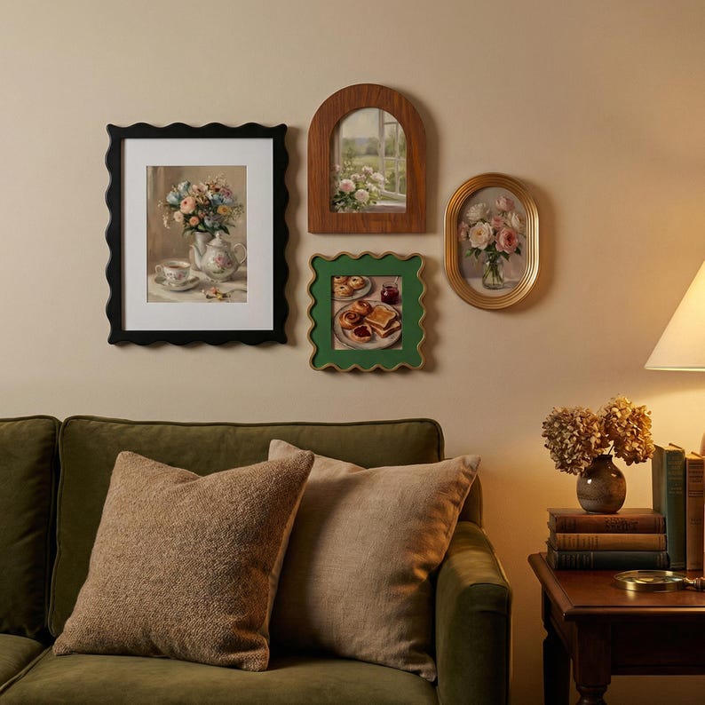 May include: A collection of four wall-mounted picture frames featuring floral and food-themed art prints. The set includes a black scalloped frame, a wooden arched frame, a green scalloped frame, and a gold oval frame, all arranged on a wall.
