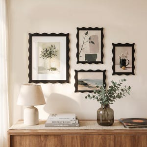 May include: A display of framed art prints with black, wavy-edged frames, featuring botanical and landscape scenes. A beige table lamp and a glass vase with greenery sit on a wooden cabinet. Books and vinyl records are also present.