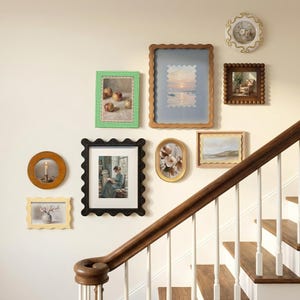 Op de afbeelding: Een verzameling ingelijste kunstwerken in verschillende vormen en maten, waaronder een ronde lijst met een kaars, een groen geruite lijst met een stilleven van appels en een grote lijst met een zeegezicht. De lijsten hangen aan een crèmekleurige muur bij een houten trap.
