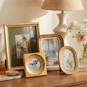 May include: A collection of four picture frames in various shapes and finishes, including a large rectangular gold frame, a square gold frame, a rounded rectangular wood frame, and an oval gold frame, displaying various art prints.