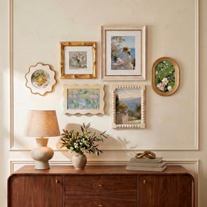 Peut inclure: Une composition murale de galerie comprenant six cadres d'art de formes et styles variés, incluant des cadres dorés, festonnés et perlés. En dessous, une lampe de table beige et un vase en céramique avec de la verdure sur un buffet en bois.