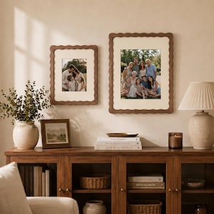 May include: Two wooden picture frames with scalloped edges and cream-colored mats, each containing a family photograph. The frames are mounted on a wall above a wooden console table, which holds books, a vase with greenery, and a table lamp.