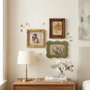 May include: A display of framed pictures and a table lamp on a wooden bedside table. The frames include a gold-framed photo, a brown beaded frame with a floral print, and a green scalloped frame with a flower painting. A vase of flowers sits on the table.