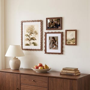 Puede incluir: Una composición de pared con tres marcos de madera de bordes ondulados y con cuentas, que muestran una lámina botánica y dos fotografías familiares. Una lámpara de mesa color crema, un frutero y tres libros apilados sobre un aparador de madera.
