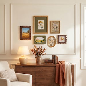 Op de afbeelding: Een galerijwand met zes ingelijste kunstwerken in verschillende vormen en maten, waaronder bloemenprints en landschappen, boven een houten dressoir met een keramische lamp, een vaas met gedroogde bladeren en enkele boeken.