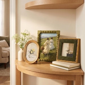 May include: A collection of three picture frames on a wooden table. One oval gold frame holds a floral print, a rectangular olive green beaded frame displays a landscape painting of two figures, and a square green velvet frame with gold beads.