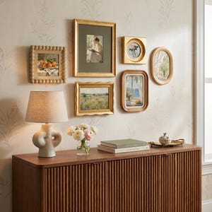 May include: A gallery wall arrangement featuring six framed art prints in gold and wood tones, a beige table lamp with a sculptural base, a glass vase with white and pink flowers, and two stacked books on a dark wood slatted sideboard.