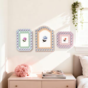 May include: Three decorative wall art pieces with floral designs. Each piece features a different flower: a purple hydrangea, a blue hydrangea, and a red rose. The frames have ornate, colorful borders with a mix of geometric and floral patterns. The art is displayed on a white wall.