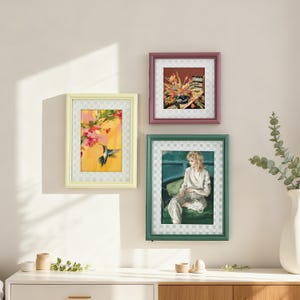 May include: Three framed art prints hang on a wall. The frames are yellow, burgundy, and green. The art includes a hummingbird, floral arrangement, and a portrait of a woman. The frames have a checkered pattern.