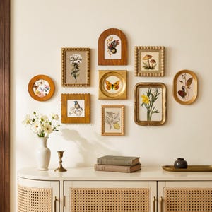 Puede incluir: Una pared de galería con nueve marcos variados en acabados de madera, oro y cuentas, que muestran ilustraciones botánicas y de insectos. Debajo, un jarrón blanco con flores blancas, un candelabro de latón y dos libros sobre un aparador.