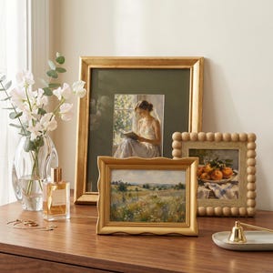 May include: A collection of three picture frames including a large gold frame with a portrait of a person reading, a smaller gold frame with a wavy edge displaying a landscape, and a square frame with a beaded border showing a bowl of oranges.