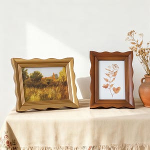 May include: Two decorative picture frames with wavy edges. One is gold-colored, displaying a landscape painting. The other is a dark wood frame, showcasing a botanical print. Both frames are on a beige tablecloth.