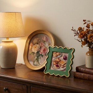 May include: A square picture frame with a wavy, scalloped edge in a deep green finish with gold trim. The frame holds an illustration of a breakfast scene featuring plates of pastries, toast, and a jar of jam, displayed on a wooden surface.
