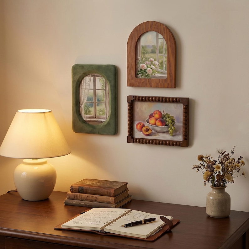 May include: Three wall-mounted picture frames featuring art prints. One frame is arched with a wood grain finish, one is rectangular with a beaded edge, and one is oval with a green velvet-like texture. The prints depict a window view and fruit.