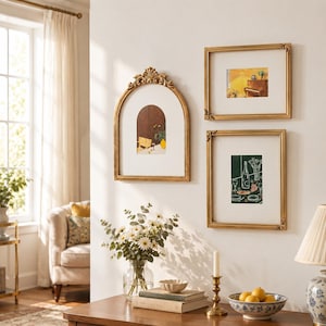 May include: Three gold-toned picture frames mounted on a wall. One frame is arched with ornate detailing at the top, while two are rectangular with corner accents. Each frame contains a white mat and a colorful illustration of interior scenes.