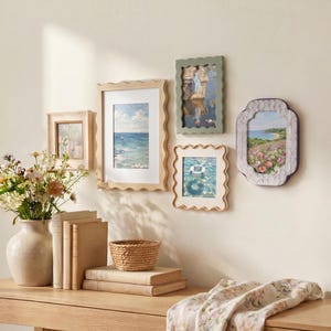 Peut inclure: Un ensemble de cinq cadres photo muraux aux bords ondulés et décoratifs, présentant diverses impressions de paysages et de nature. Les cadres sont disposés en groupe, avec des finitions en bois clair, vert sauge et violet texturé.