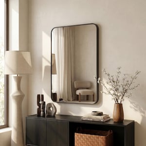 Pode incluir: Um espelho de parede retangular com cantos arredondados e uma moldura fina e escura. O espelho reflete um quarto com cortinas brancas e uma cadeira. Está posicionado sobre um aparador de madeira escura com candeeiro, objetos e vaso.