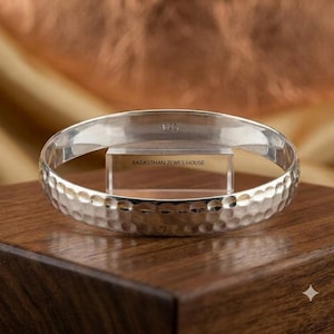 Puede incluir: Un brazalete de plata con una textura martillada en la superficie exterior. El interior de la banda es liso y presenta el texto grabado 925. Un soporte de acrílico transparente dentro del brazalete muestra el texto RAJASTHAN JEWES HOUSE.