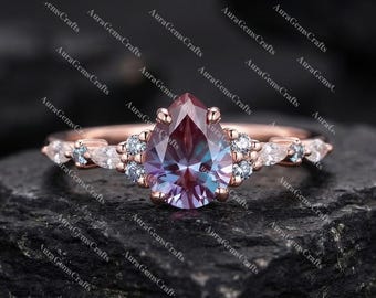 Puede incluir: Anillo de oro rosa con una piedra preciosa en forma de pera que cambia de color. El anillo está adornado con pequeñas piedras preciosas redondas y en forma de marquesa en tonos azules y blancos. El anillo está sobre un fondo oscuro y texturizado.