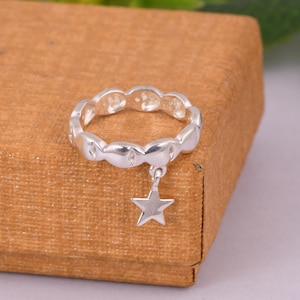 May include: A silver ring with a repeating oval design and a dangling star charm. The ring is displayed on a brown surface, showcasing its polished finish and delicate details. This piece of jewelry is a charming accessory.