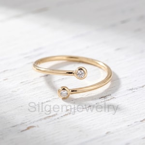May include: A gold open-style ring featuring two circular ends, each set with a single small clear gemstone. The band is polished and smooth, with the text Silgemjewelry visible in the foreground. The design is simple and circular in shape.