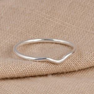 May include: A silver ring with a curved, wave-like design. The ring is slender and has a polished finish. The ring is displayed on a neutral-coloured fabric background.