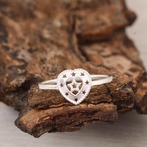 Könnte beinhalten: Silberner Ring mit herzförmigem Design, verziert mit Sternaussparungen und einem zentralen Stern. Der Ring wird vor einem natürlichen Holzuntergrund präsentiert, der die filigrane Handwerkskunst des Schmucks hervorhebt.