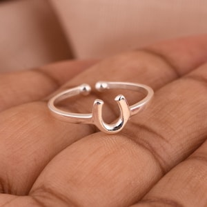 Könnte beinhalten: Ein silberfarbener Hufeisenring. Der Ring hat ein Hufeisen-Design in der Mitte mit einer glatten, polierten Oberfläche. Der Ring ist verstellbar und sitzt auf der Hand einer Person.