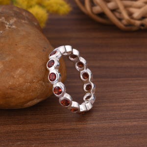 Op de afbeelding: Een zilveren ring met een doorlopende rij ronde, rode edelstenen. De ring wordt gepresenteerd tegen een natuurlijke achtergrond met een bruine steen en een houten oppervlak, waarbij het ontwerp en de kleur van het sieraad worden benadrukt.